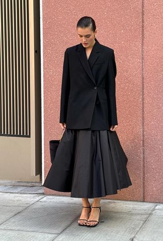 A woman wearing a black blazer with a black full skirt, black strappy sandals, and a black handbag.