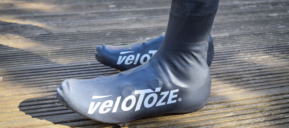 A pair of feet on a wooden deck wearing black veloToze overshoes with white logos