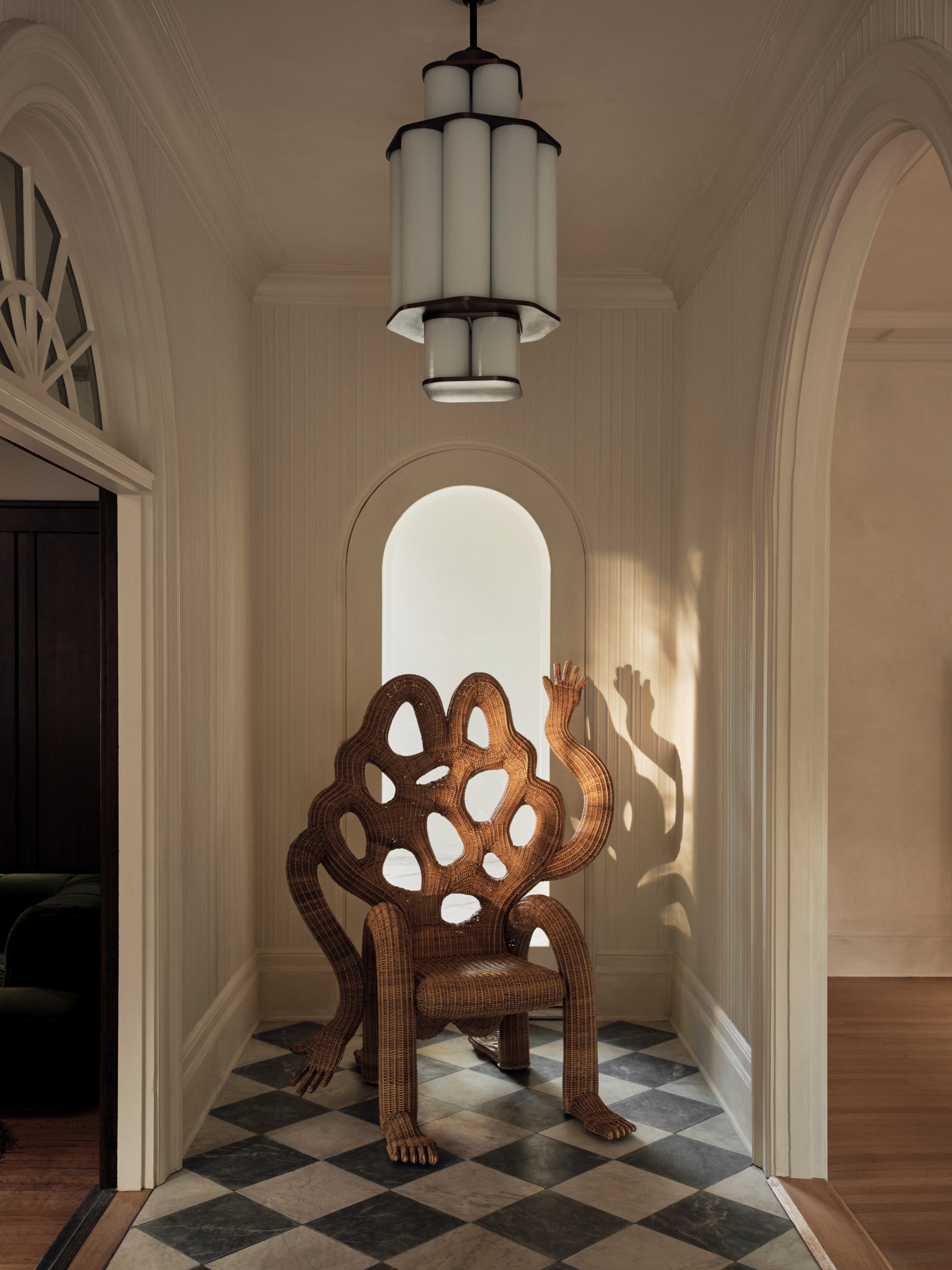A whimsical wicker chair, sitting in a sun-lit entryway with checkered white and black floors and a pendant chandelier, with two hands and a perforated back, with four feet.