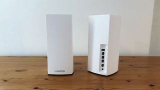 Två stycken Linksys Velop WiFi 6 AX4200 står placerade på ett träbord mot en vit bakgrund.