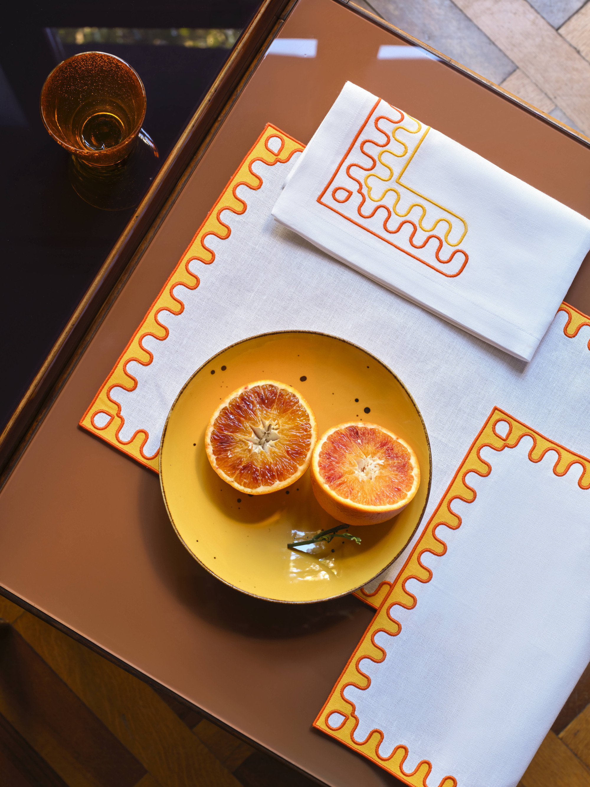 Image from above of white placemates and napkins that have a bright orange and pink wavy boarder. There is an orange plate with a grapfruit on it.
