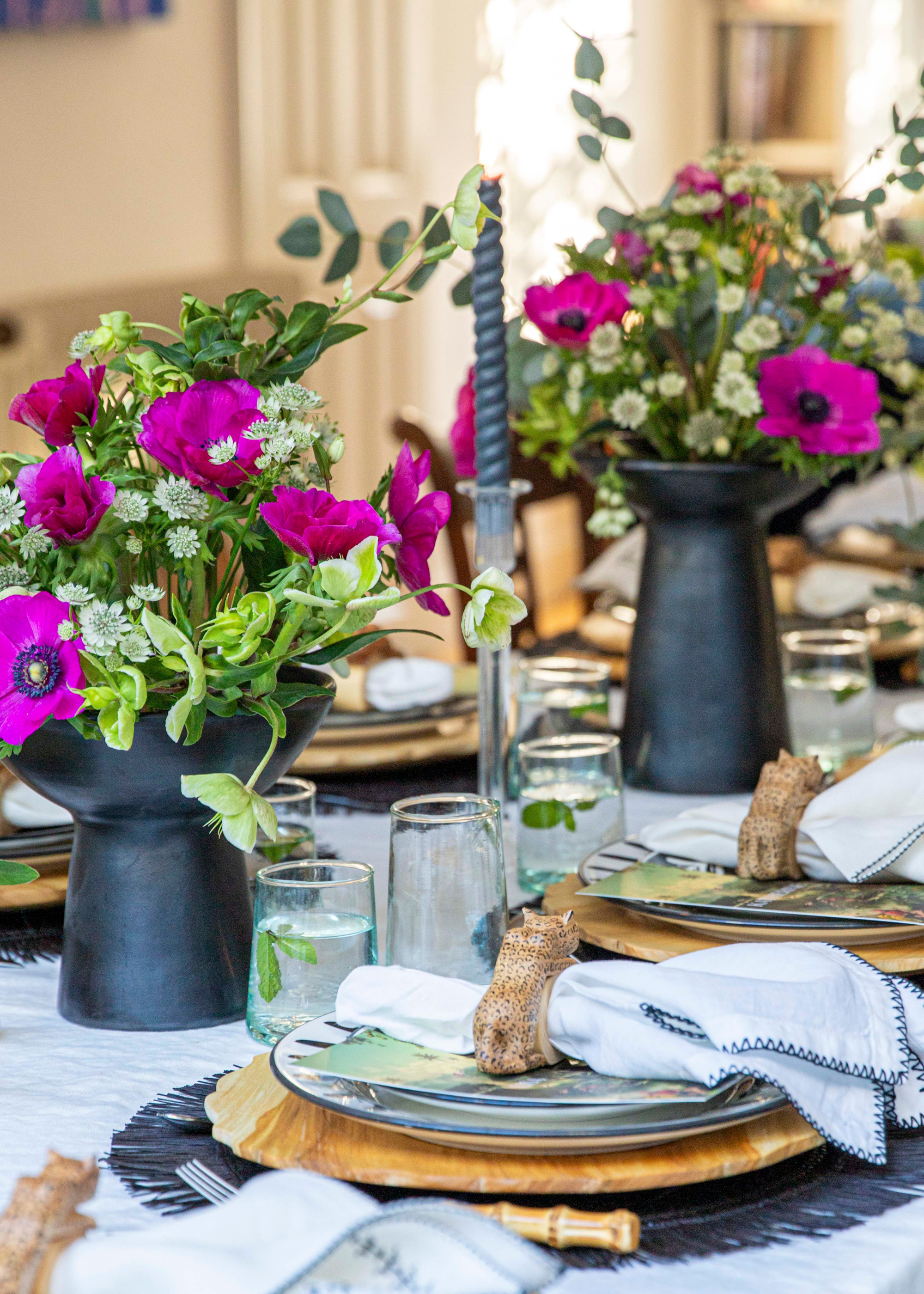 A dining table set with plates, napkins, glasses, floral arrangements in black vases, and tapered candles
