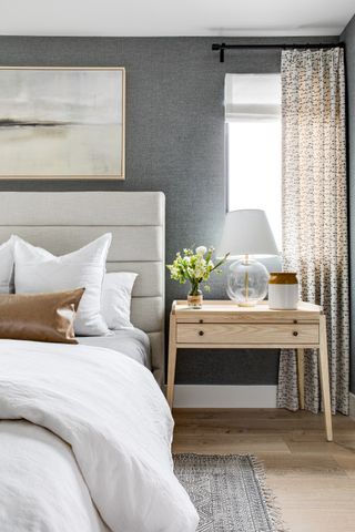 grey and white bedroom with grey textured wall covering, pale grey upholstered bed, pale blond wood side table, graphic print drapes, white bedlinen, artwork