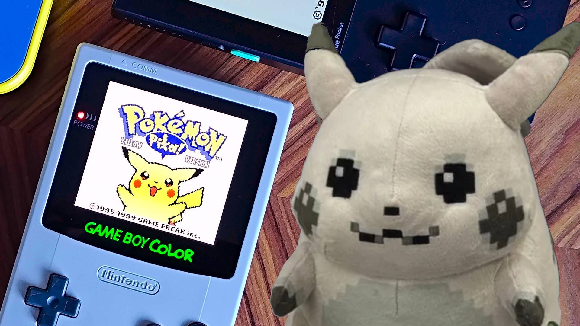 A Game Boy Color with a sepia Pikachu plushie beside it