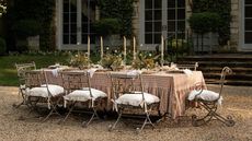 large long outdoor table styled on a shingle patio area with a pink plaid ruffle tablecloth, ornate chairs, laid with flowers and tall taper candles