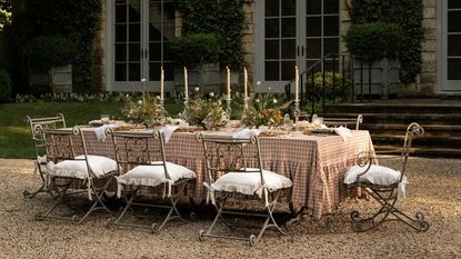 large long outdoor table styled on a shingle patio area with a pink plaid ruffle tablecloth, ornate chairs, laid with flowers and tall taper candles