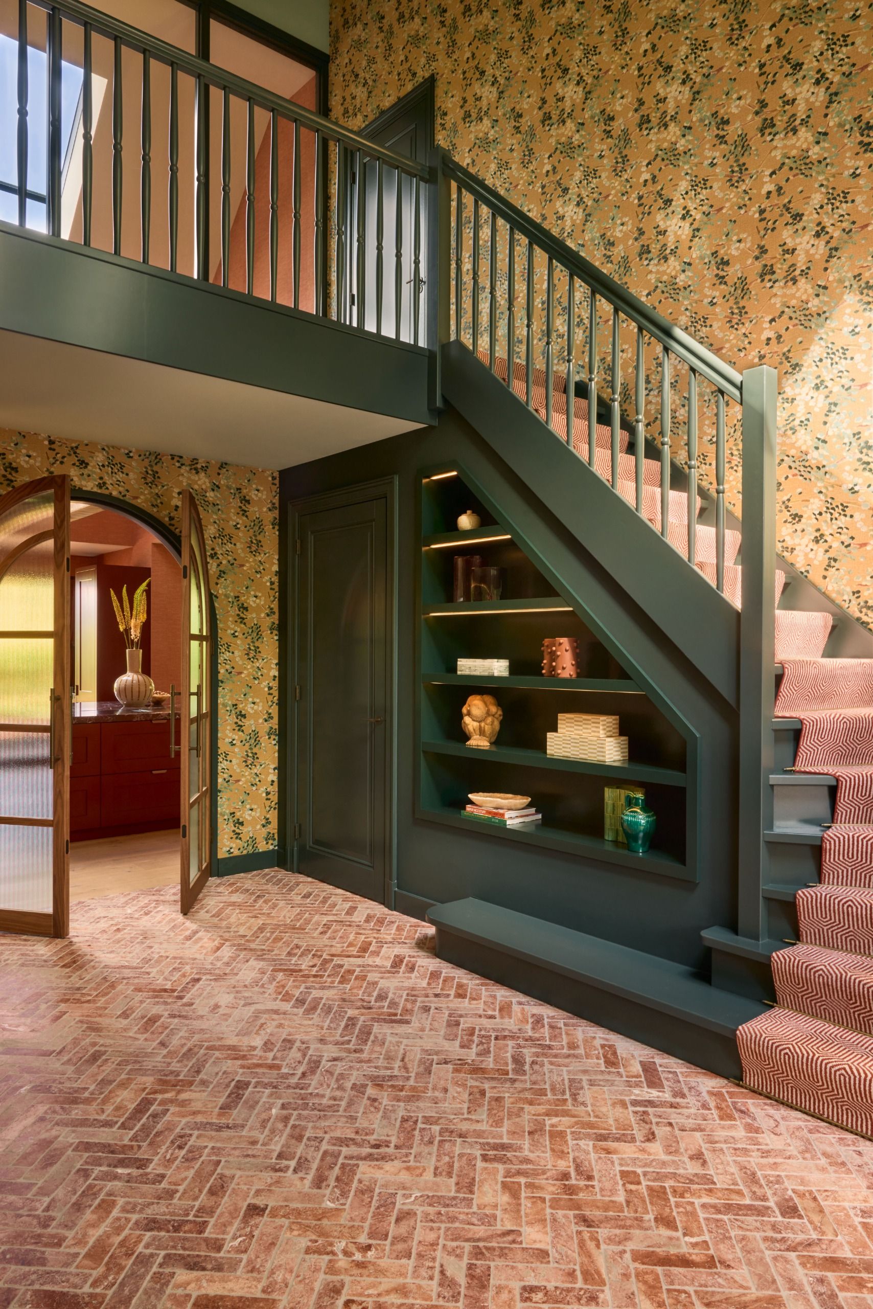 A grand entryway with a large green staircase with a pink runner, yellow floral print wallpaper, and parque brick flooring.