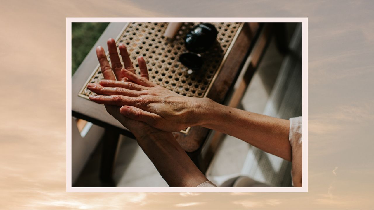 A close up of a woman applying moisturiser to her hands, whilst wearing a pink top and sitting besides a wooden table with cosmetics on it/ in a cream and purple sunset-style template