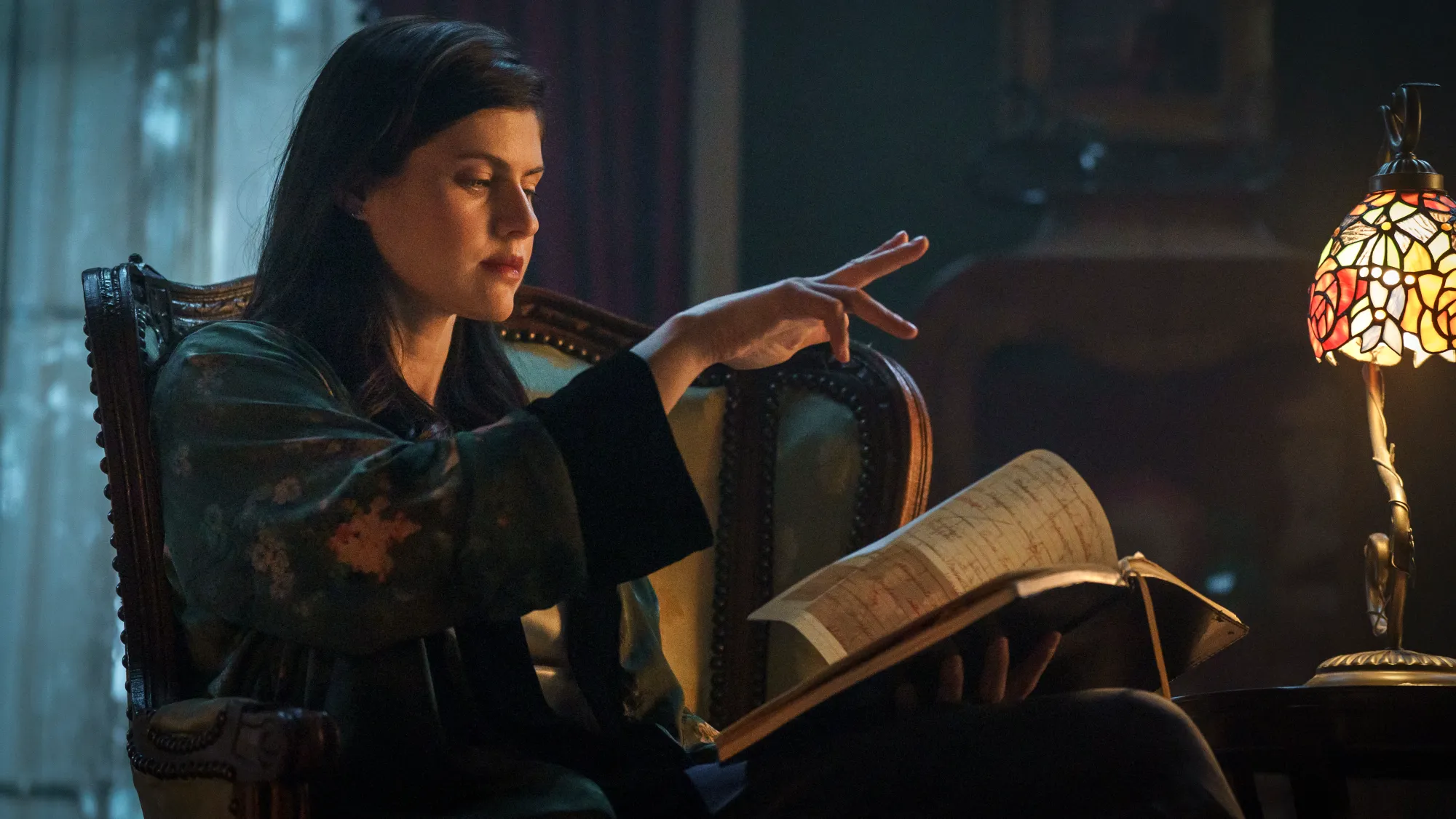 alexandra daddario opening a spellbook and sitting at a desk lamp in the mayfair witches