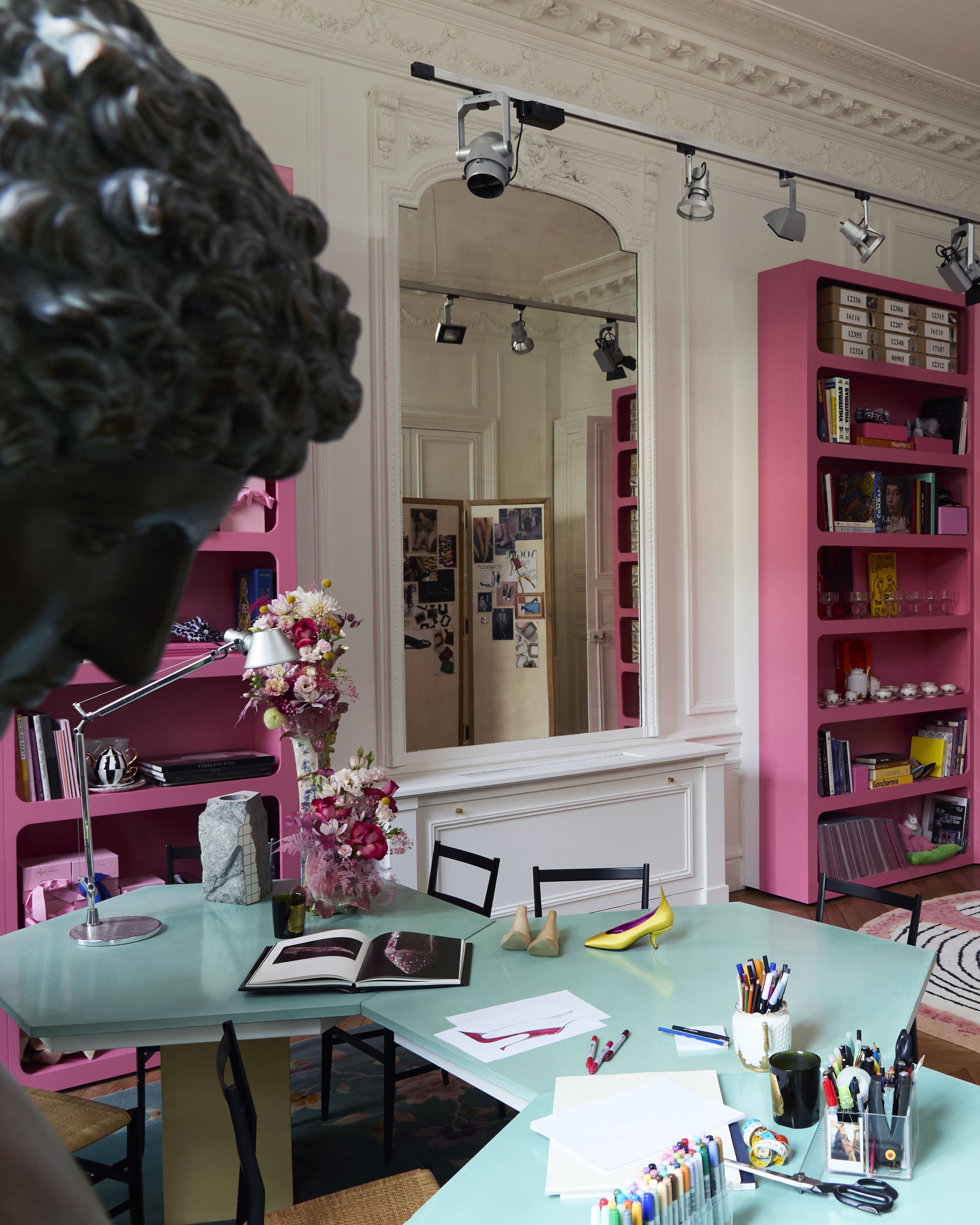 A colorful office studio with a sweeping, aquamarine green desk filled with stationery, books, and shoes, bright pink bookshelves embracing it, a bronze sculpture of a boy, and abstract tufted rugs in white, black, and pink.