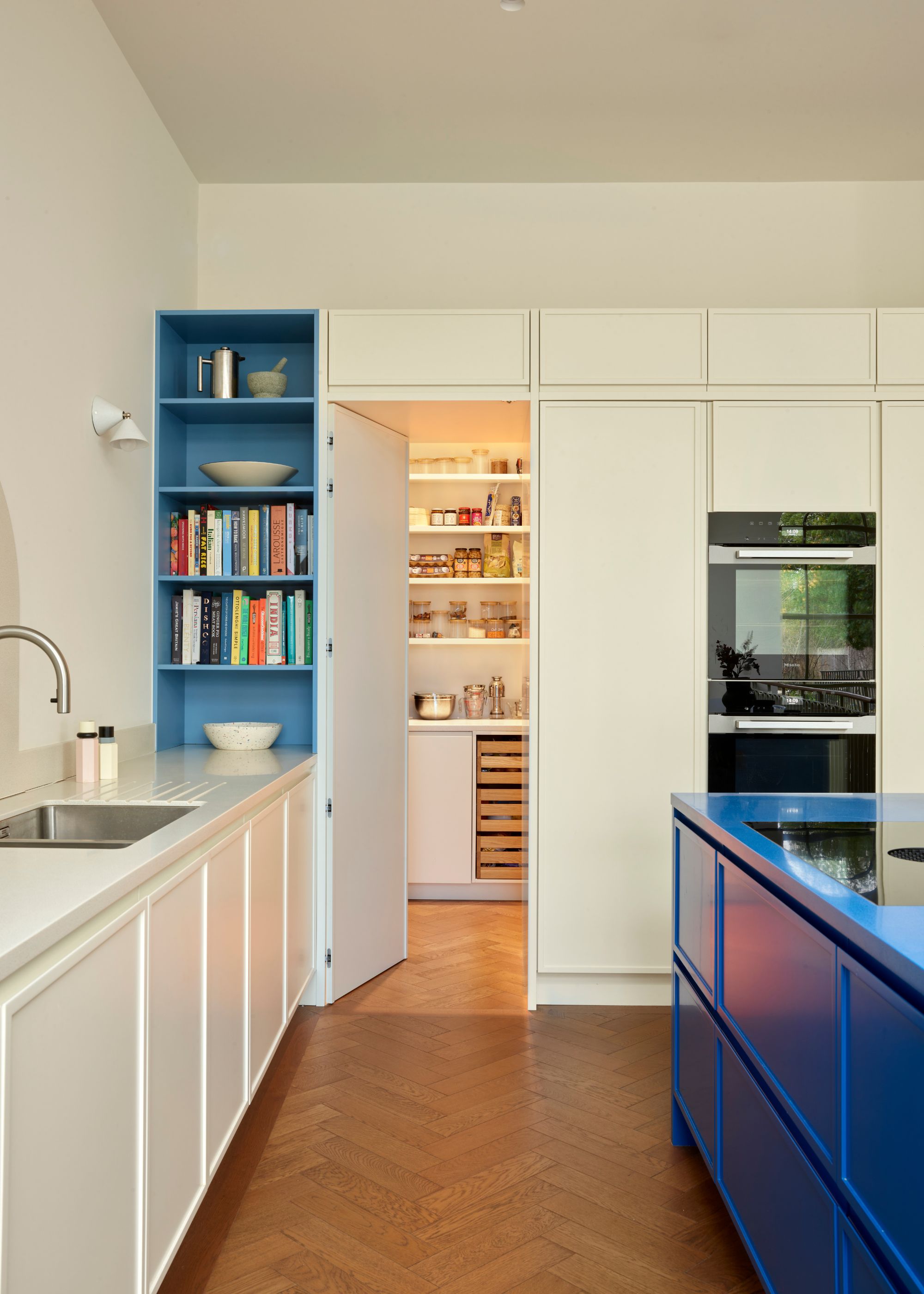 A modern, minimalist kitchen with off-white cabinets, parquet flooring, a cobalt blue kitchen island, and a pop of blue paint on the corner cabinet with open shelving.
