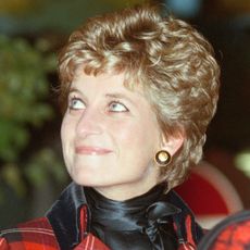 A headshot of Princess Diana wearing a red-and-black outfit and big black and gold Chanel button earrings looking to the left with a smile