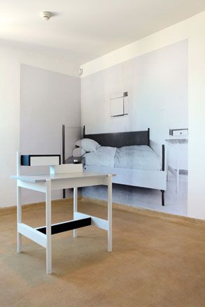 A room with white table in the middle of the room on brown floor and a wall paper featuring a bed