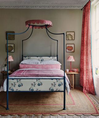 Bedroom space with khaki painted walls, metal four poster bed frame with scalloped, pink patterned bunting and upholstered head and footboard. Pink patterned and white linen and cushions, red patterned curtains, textured natural rug with scalloped edging and red stripe, two matching wooden side tables with matching red lampshades, framed pictures and art on the wall