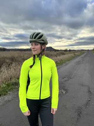 A woman in a bright fluorescent jacket looking to the side with a muddy field behind and a green helmet on