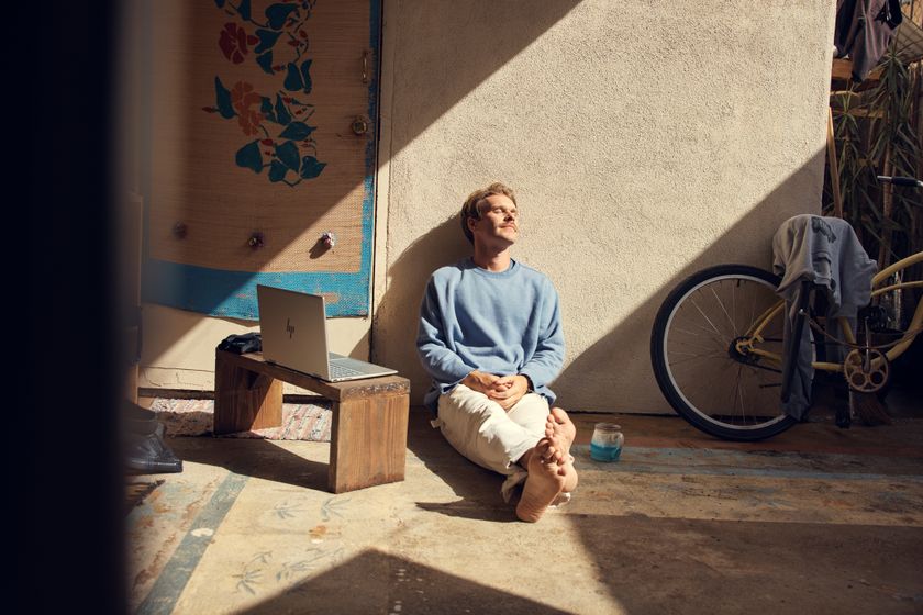 A man sat against a wall basking in the sun, with an HP laptop sat on a small bench to his immediate right. He has his eyes shut and is sat in a warm, urban space.