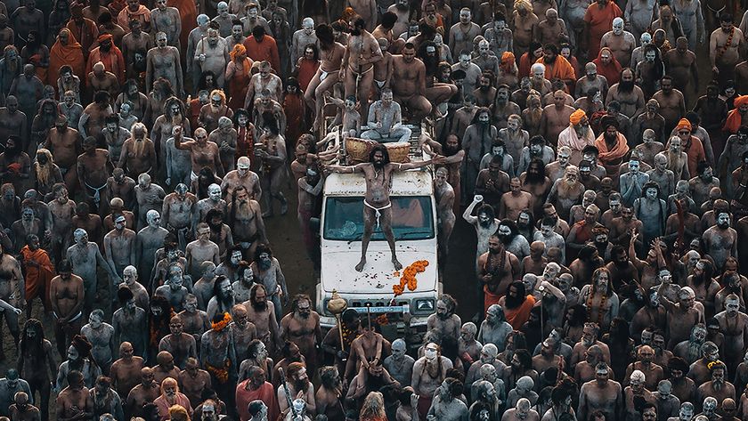 Aerial view of a large congregation of people covered in ash and paint, grouped around a vehicle, engaged in a vibrant spiritual gathering