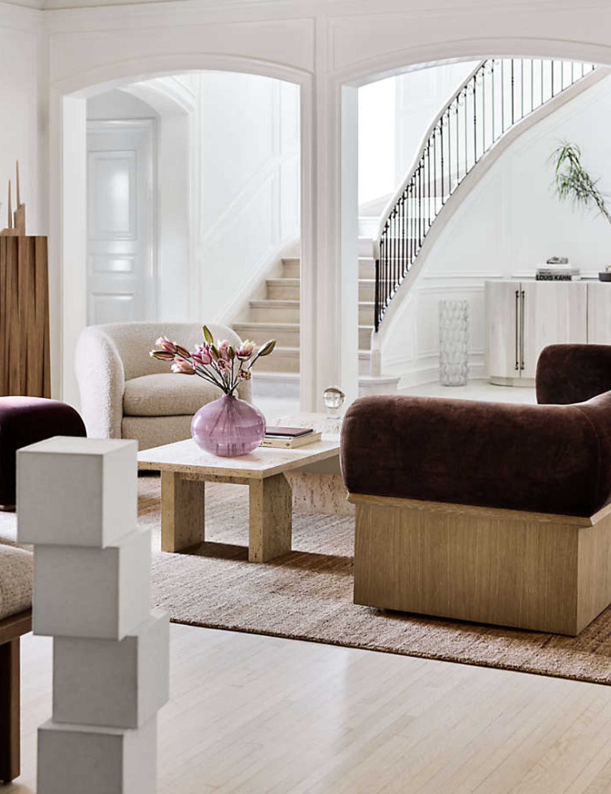 Brigette Romanek x Crate&Barrel collection, including a geometrically stacked lamp, curved sofa, and sleek travertine coffee table, photographed in a light, bright living room