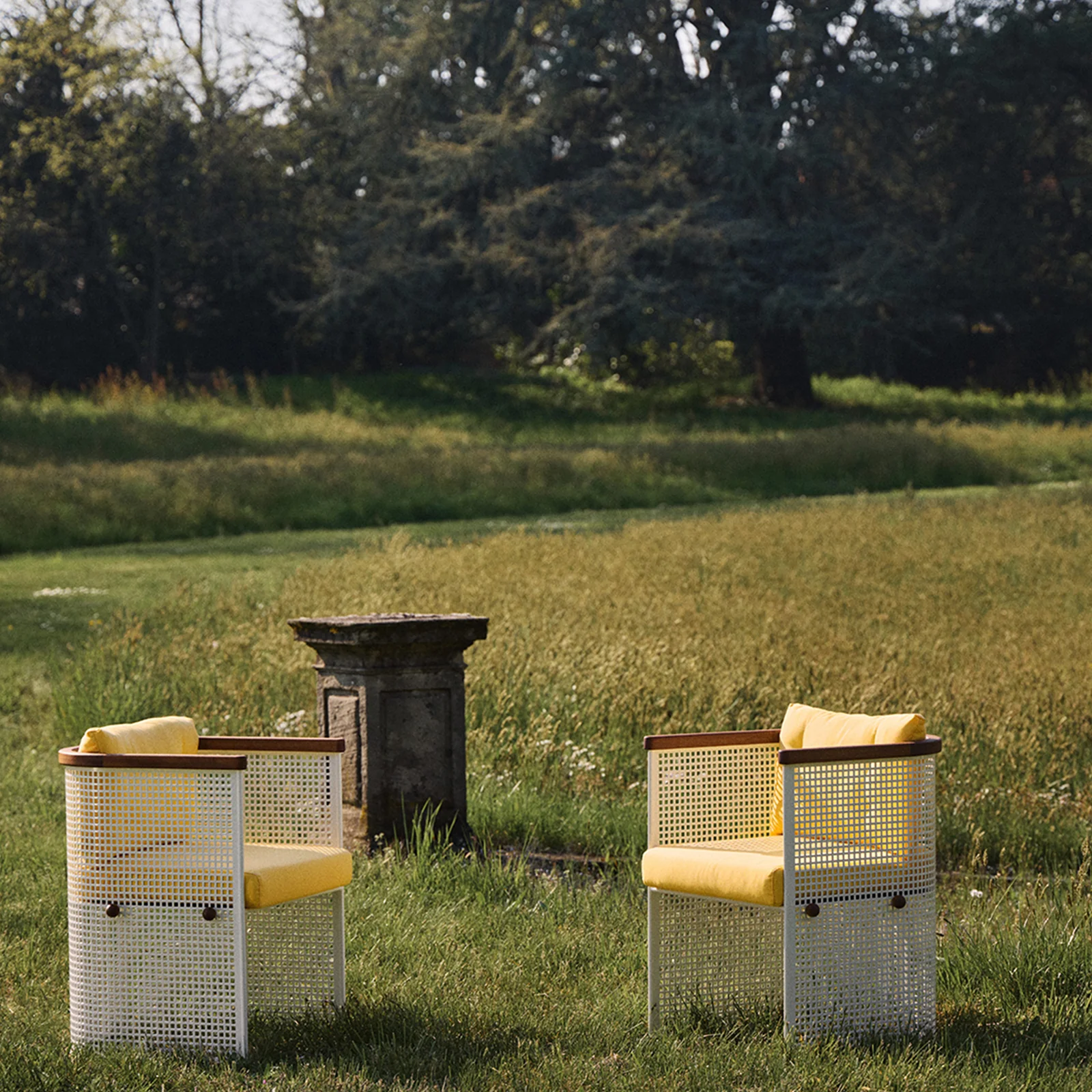 two white perforated metal chairs with yellow cushions in a meadow by a fountain