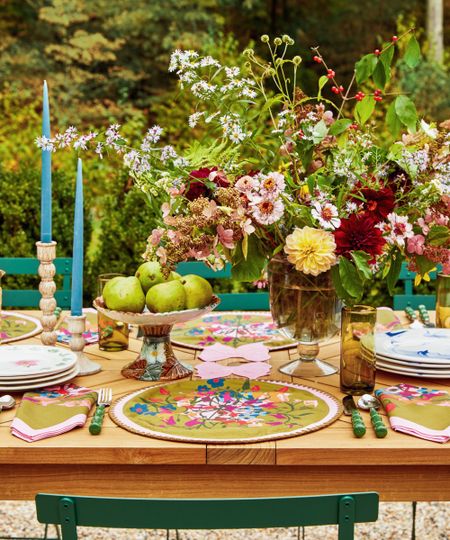 Garden with wooden dining table, green wrought iron chairs, candlesticks with bleu taper candles, white ice buckets holding wine, a floral tablecloth and a large bouquet of flowers