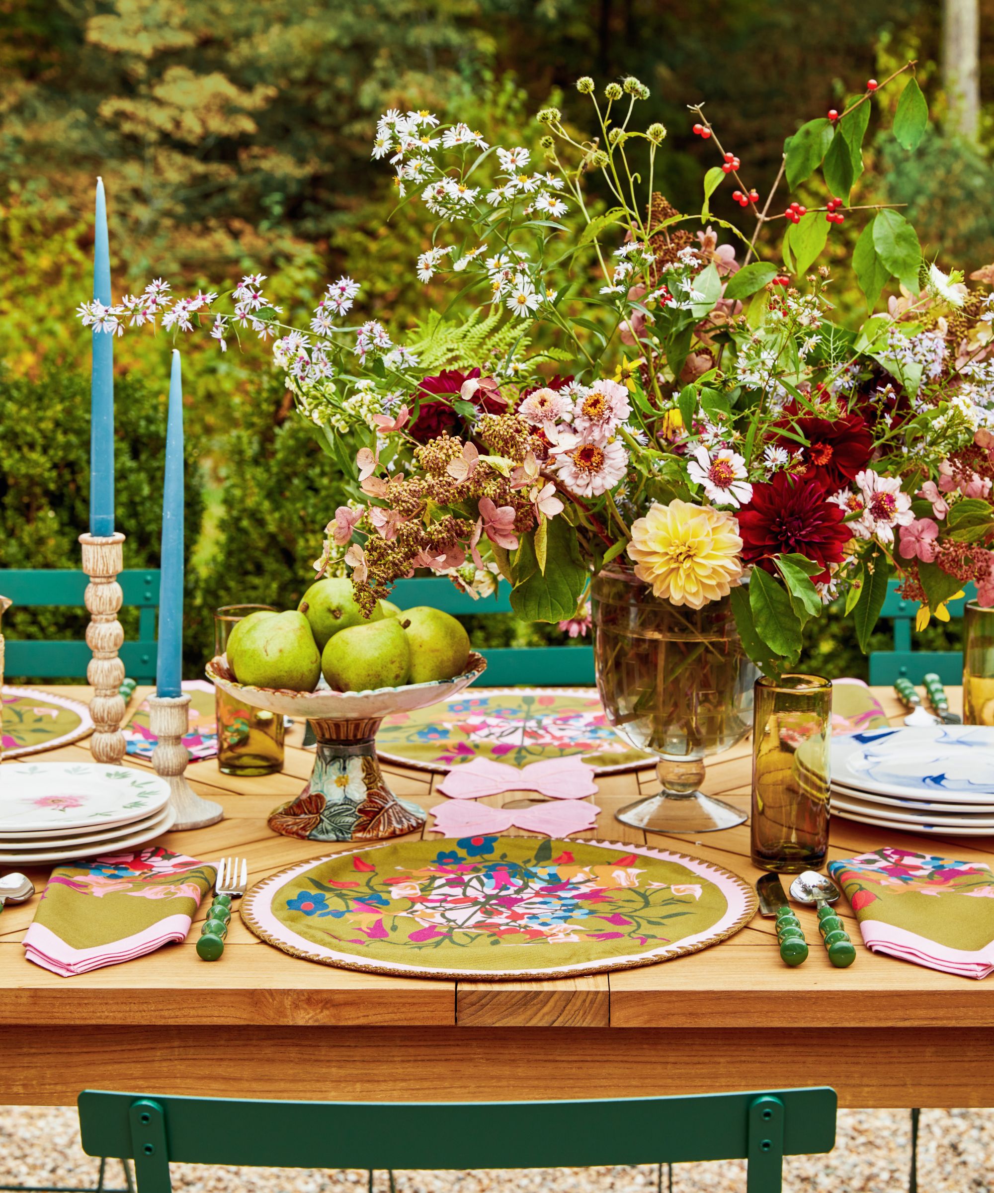 Garden with wooden dining table, green wrought iron chairs, candlesticks with bleu taper candles, white ice buckets holding wine, a floral tablecloth and a large bouquet of flowers