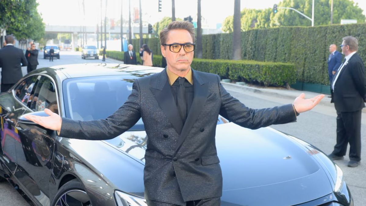 Robert Downey Jr., wearing a black suit and sunglasses, stands in front of a car with his arms in the air, in a trailer for Downey&#039;s Dream Cars shown at the WBD Upfronts 2023