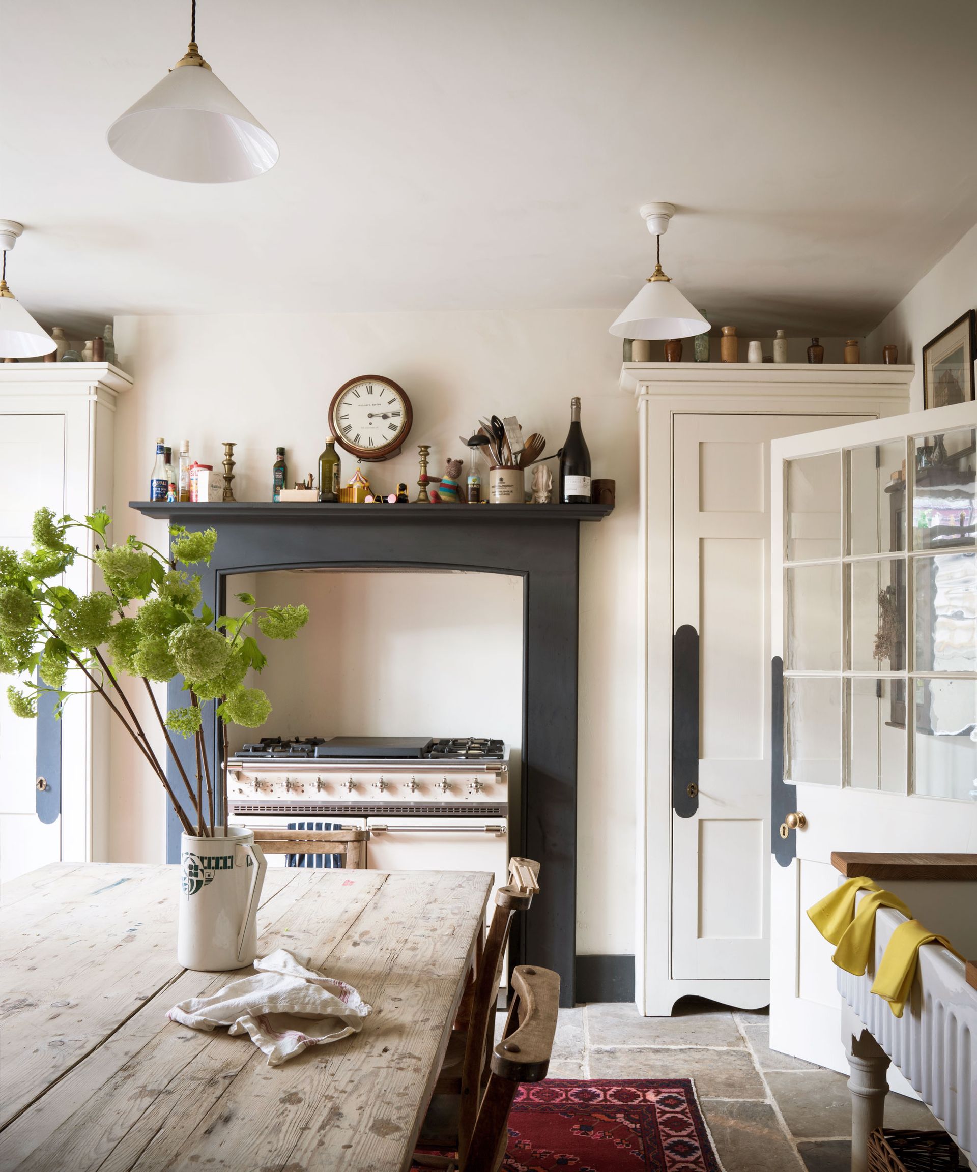 County kitchen with stone floor, large cooking range, wooden dining table, cone pendant lights, red rug on floor, white wooden cabinetry
