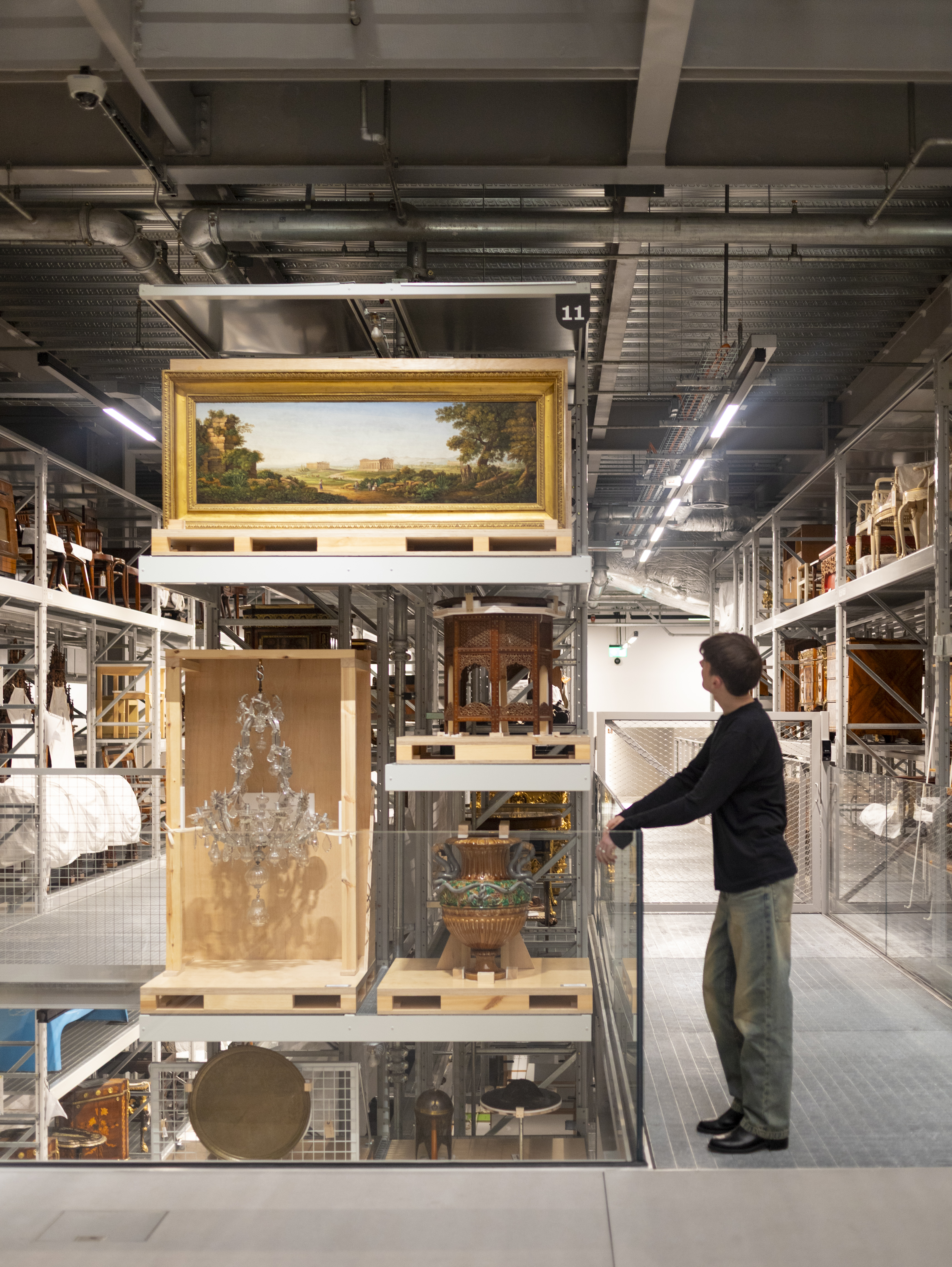 A young man dressed in a black jumper and denim jeans stares at a series of neoclassical artifacts stored in a warehouse's shelf.
