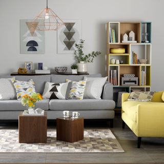 a grey living room with a grey couch with cushions with a yellow and grey geometric pattern, with two wooden cube coffee tables and a yellow armchair, brown, and a white, yellow and blue bookcase in different size rectangles