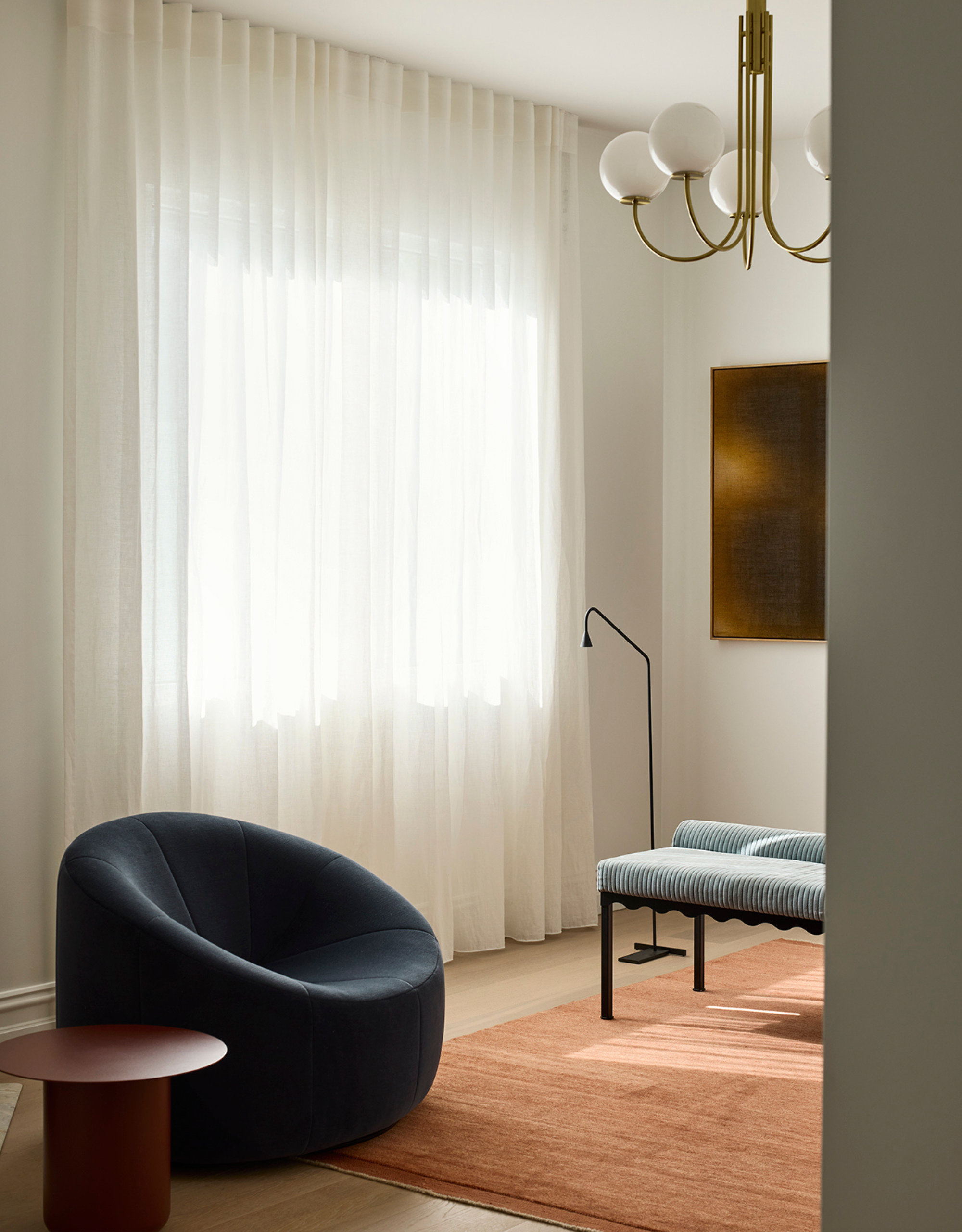 A white living room with sheer curtains, a blue sofa chair, a red side table, a striped bench, a floor lamp, a peach rug, and a statement light