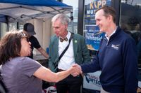 a smiling man in a blue sweater shakes hands with a woman while an older fellow in a bow tie looks on