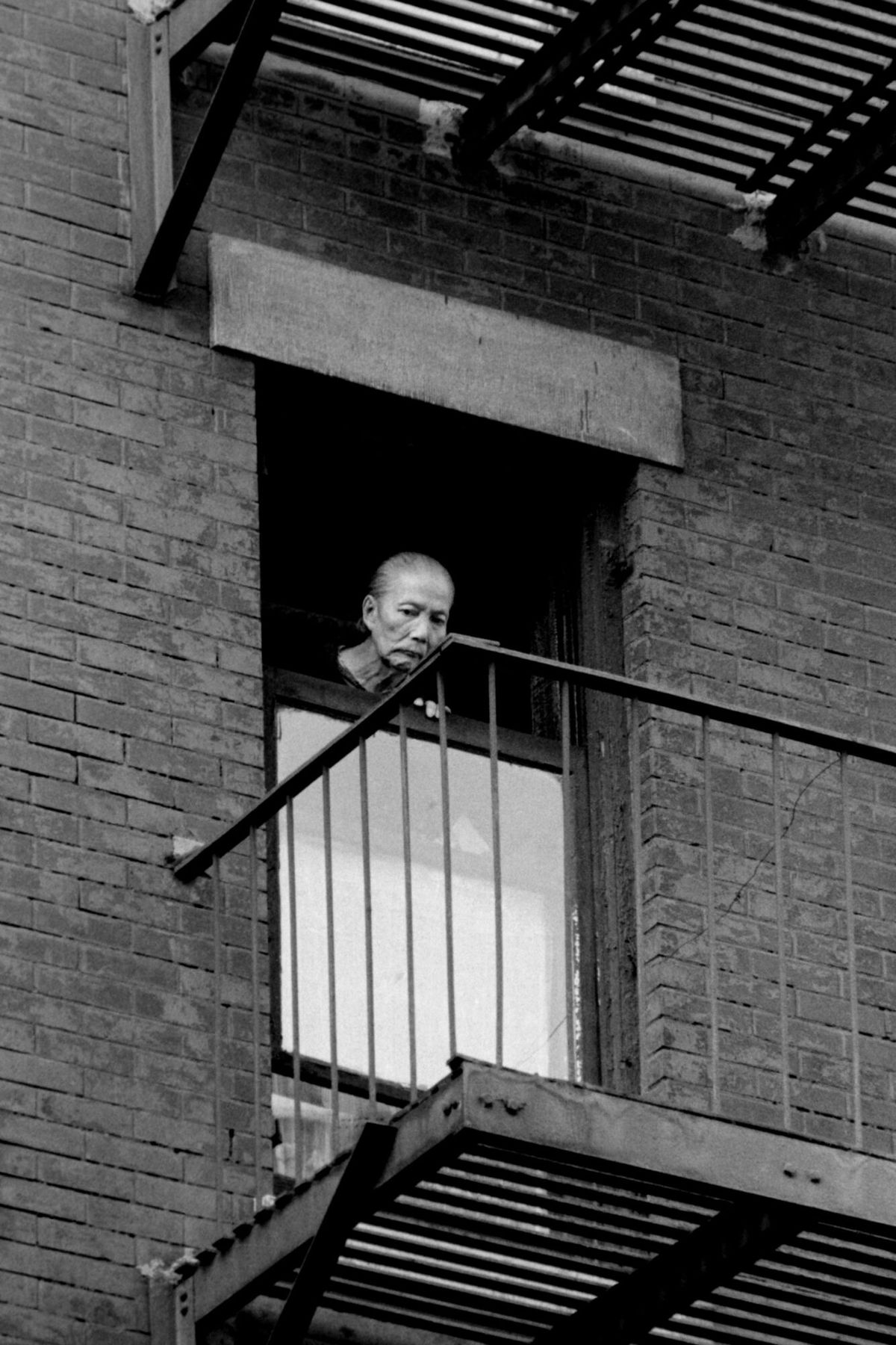 An elderly woman with her hair pulled back leans out of a brick apartment window onto the railing of a fire escape, looking down towards the street.