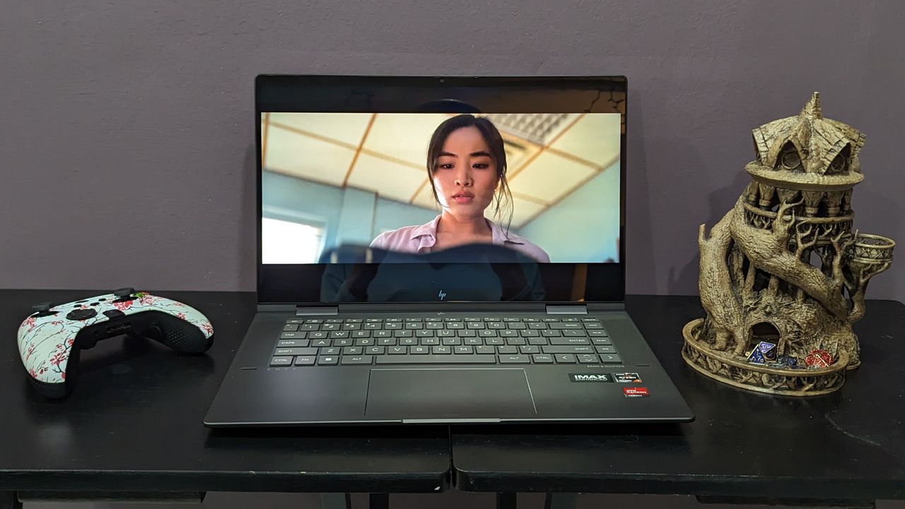 HP Envy x360 open on a black desk with a white gaming controller and a dice tower on either side of it.