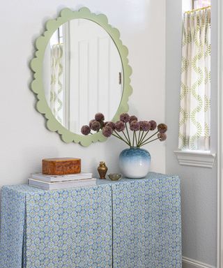 corner of room with white walls, green decorative oval shaped mirror on wall, side table with light blue patterned tablecloth