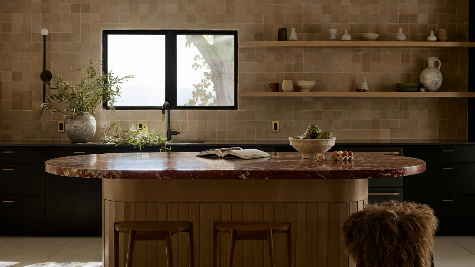 A warm neutral kitchen with tiled walls, black cabinetry, and a curved wooden island topped with a burgundy marble countertop
