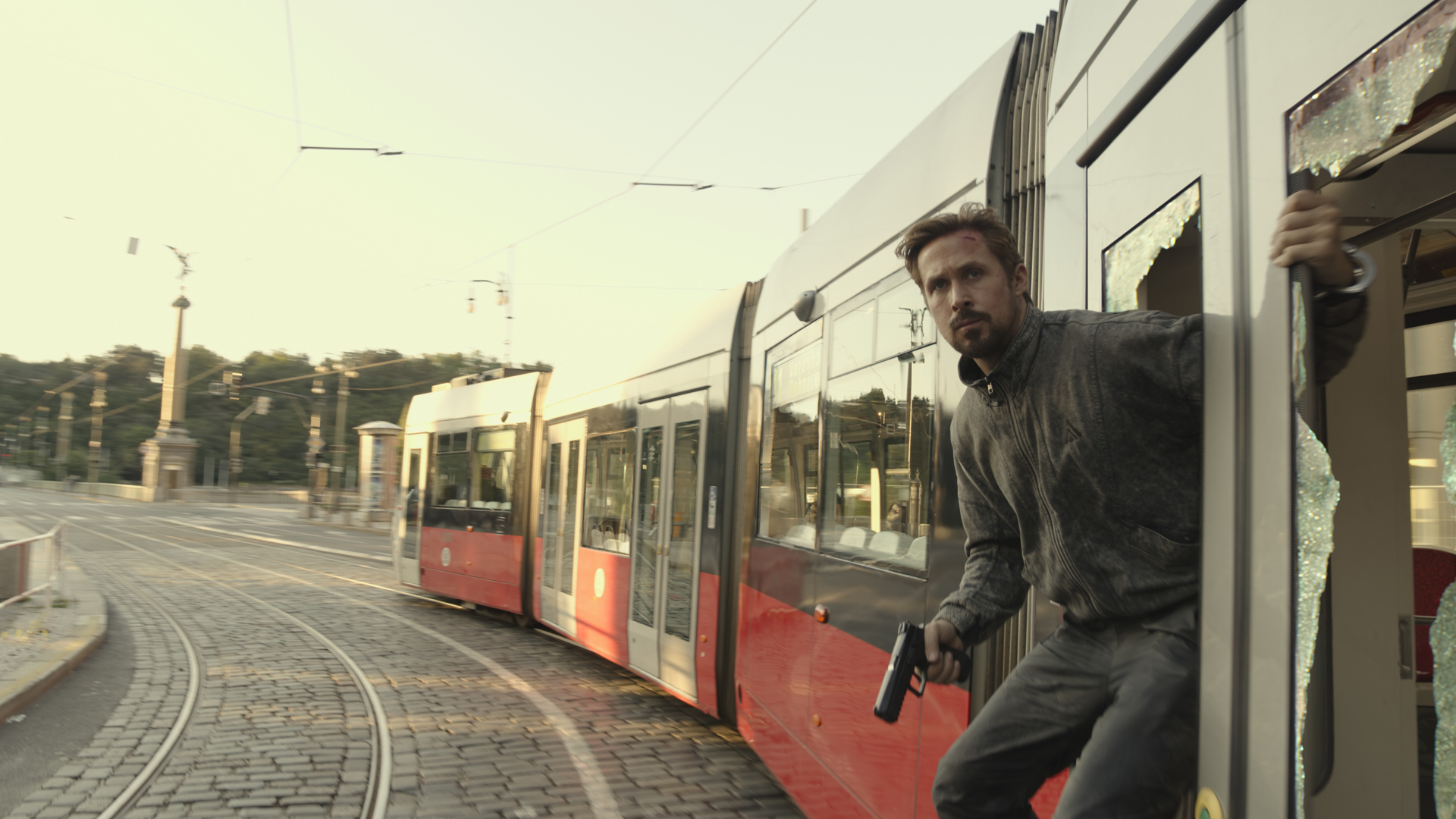 Ryan Gosling's Sierra Six hanging from the side of a train, gun in hand, in The Gray Man