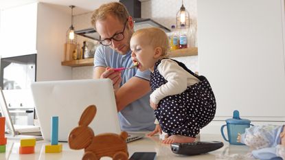 A dad working at home feeds a toddler at the same time.