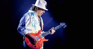 Carlos Santana wears a white wide-brimmed hat and white patterened shirt as he plays his signature salmon-colored PRS signature guitar.