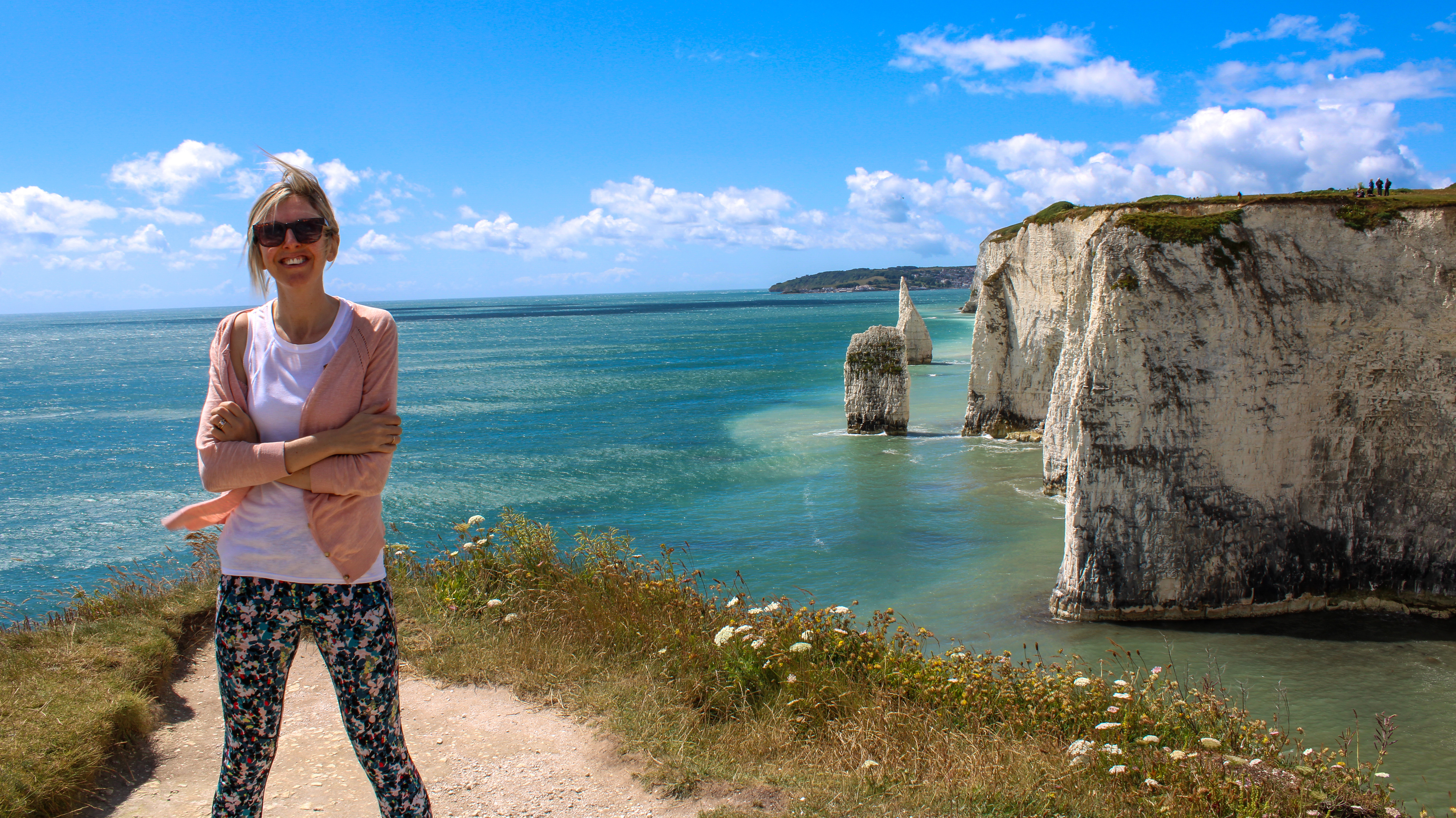 Kathryn Williams is on a coastal path, standing on a cliff with the ocean behind her. She wears leggings, a t-shirt and a long cardigan with sunglasses.