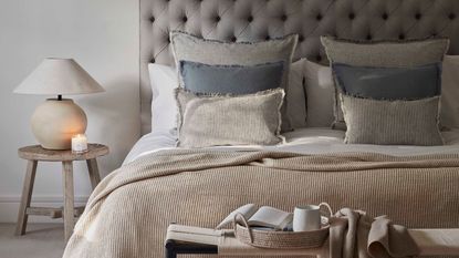 A cozy bed in neutral colors with a large table lamp beside it, and a bench with a cup of coffee and an open magazine
