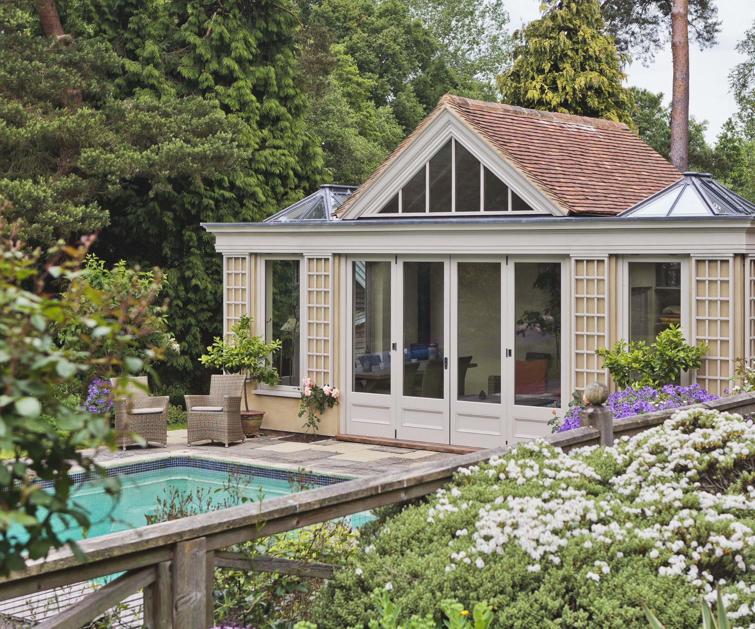 A grand cream-painted orangery-style pool house with a clay tile and glazed roof, tall French doors, trellis detailing and window boxes, surrounded by mature woodland and white flowering shrubs.