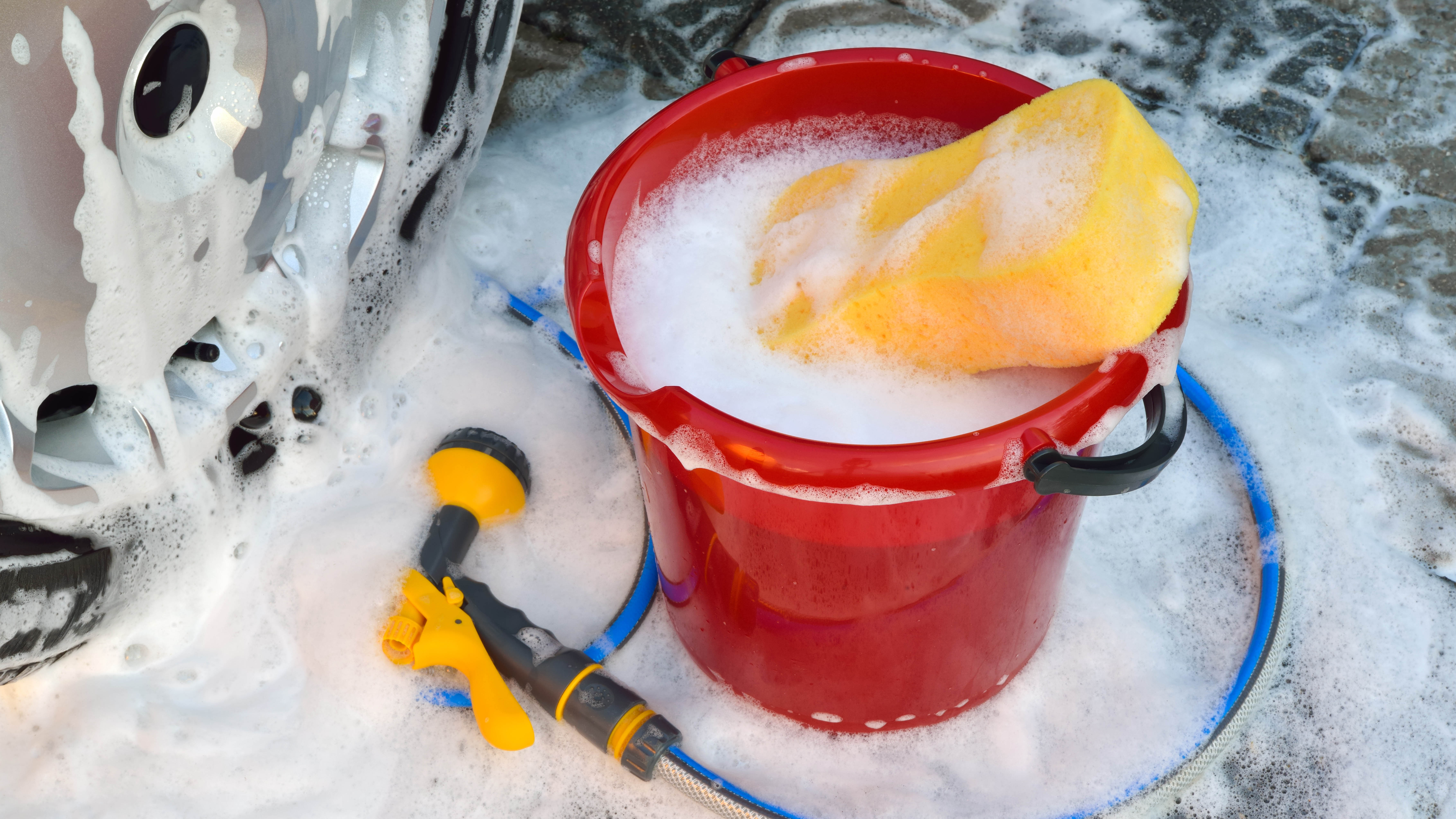 A red bucket filled with soapy water and a sponge next to a car