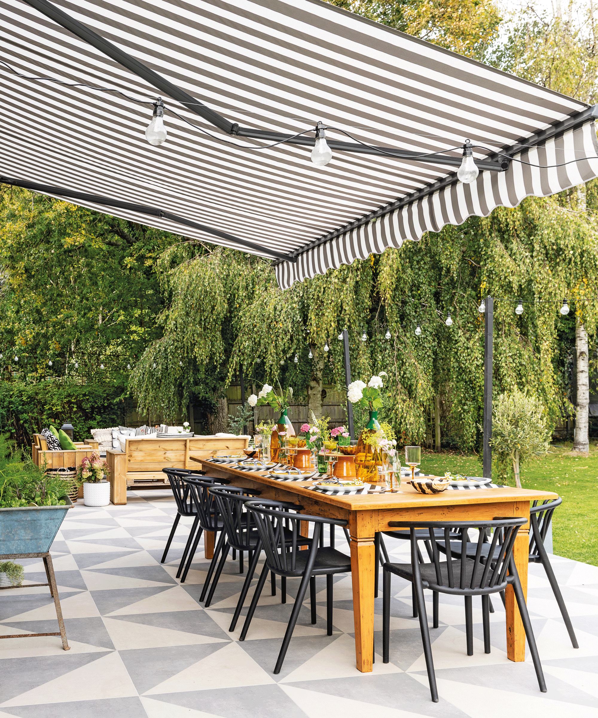 Alfresco dining set-up with fresh flowers on table top, festoon lights, striped awning shade, and large scale mono geo floor tiles.