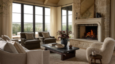 A carpeted living room with large floor-to-ceiling windows and a fire roaring in a stone fireplace with an ornate mirror resting on the mantle above. Cozily upholstered accents chairs, a sofa, and low slung charcoal coffee table holding collected objects sit before it.