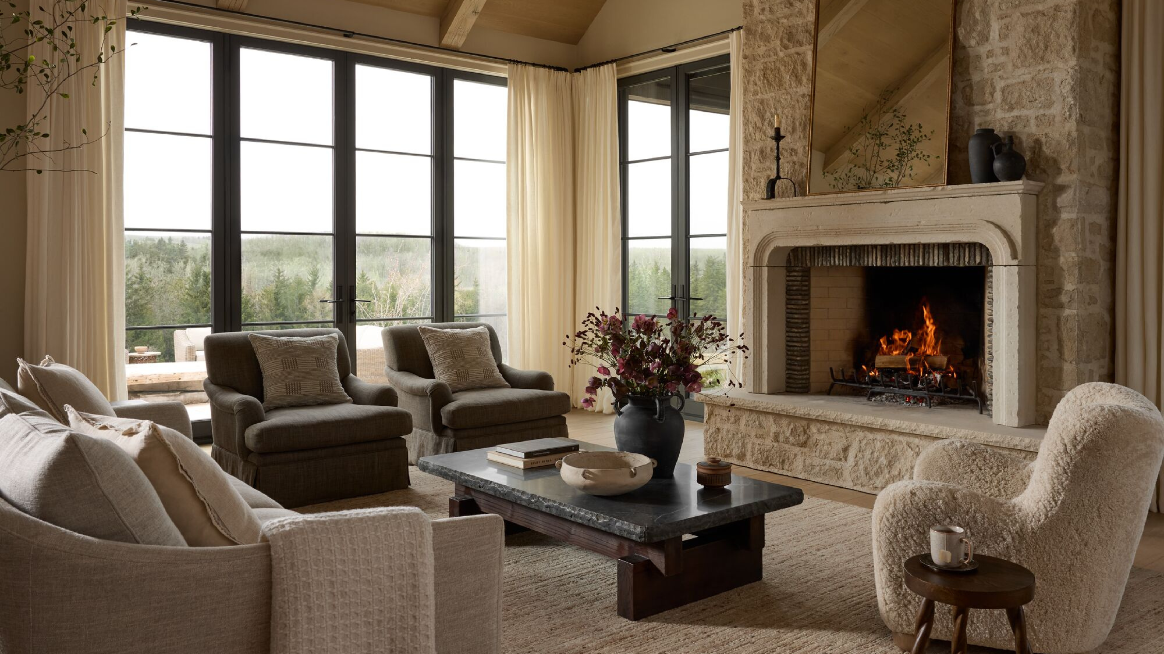A carpeted living room with large floor-to-ceiling windows and a fire roaring in a stone fireplace with an ornate mirror resting on the mantle above. Cozily upholstered accents chairs, a sofa, and low slung charcoal coffee table holding collected objects sit before it. 