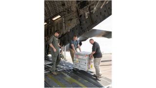 three workers carry a silver crate off the back of a cargo plane.