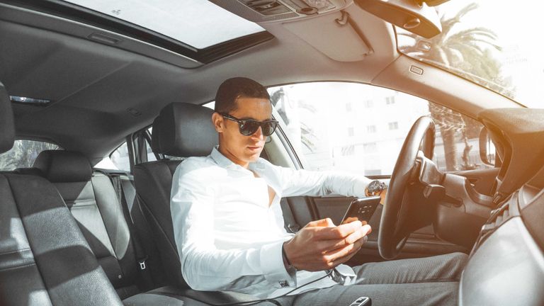 A man sitting in a car wearing the best driving sunglasses and looking at his phone
