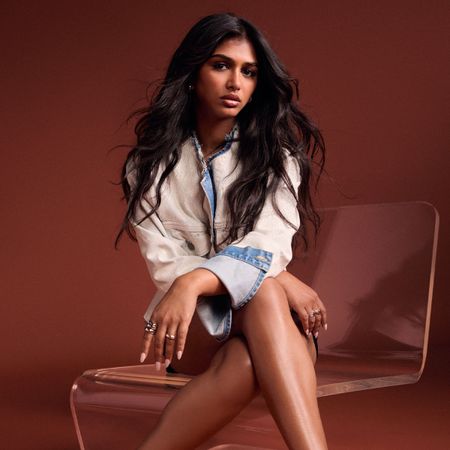 Priyanka Kedia sits on a plexiglass chair in front of a rust colored backdrop. She is wearing a white denim jacket. 