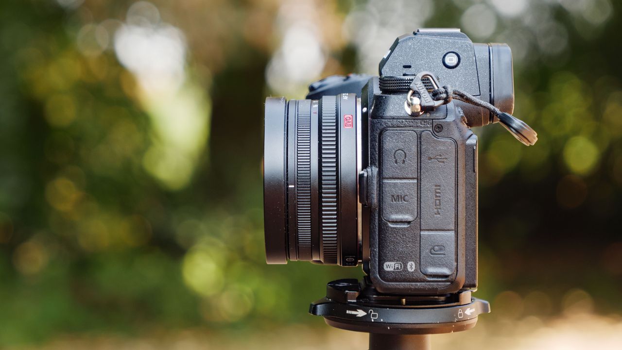 Side profile of the Viltrox AF 26mm F2.8 EVO 'pancake' lens attached to a Nikon mirrorless camera, there's dappled light through a tree behind it