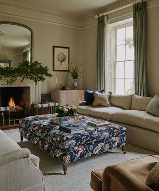 traditional neutral english living room with a large fireplace decorated with foliage and a large navy patterned ottoman